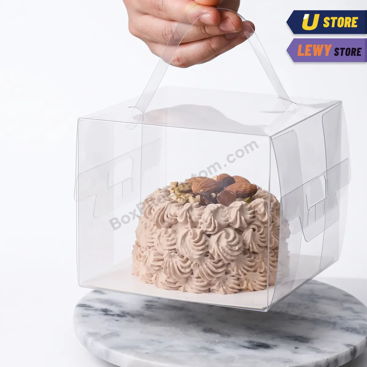 Kotak kue mika bening beralas putih berisi kue krim cokelat dan almond, di atas cake stand marmer