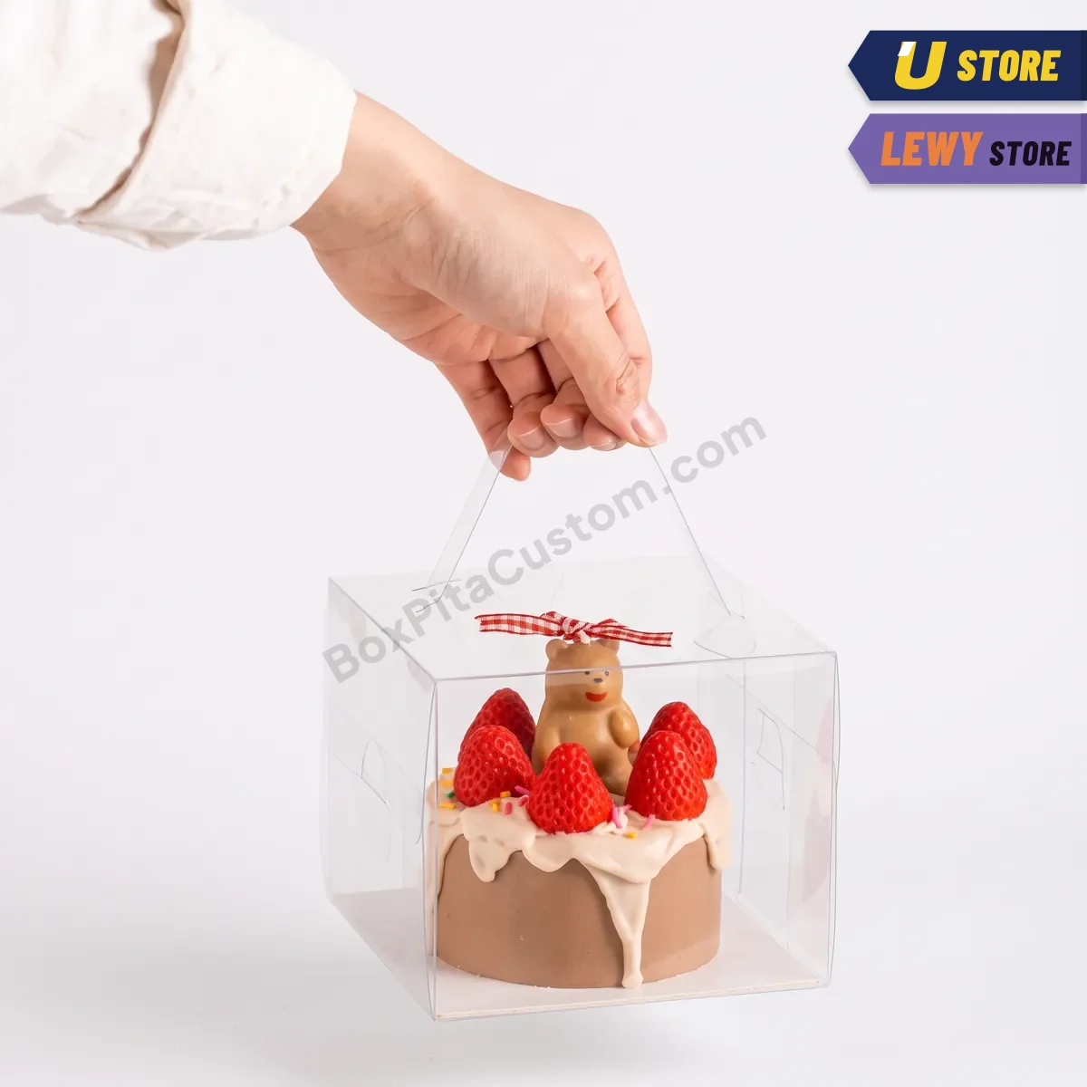 Kotak kue mika bening beralas putih ditenteng pada pegangannya, berisi kue cokelat berhias stroberi