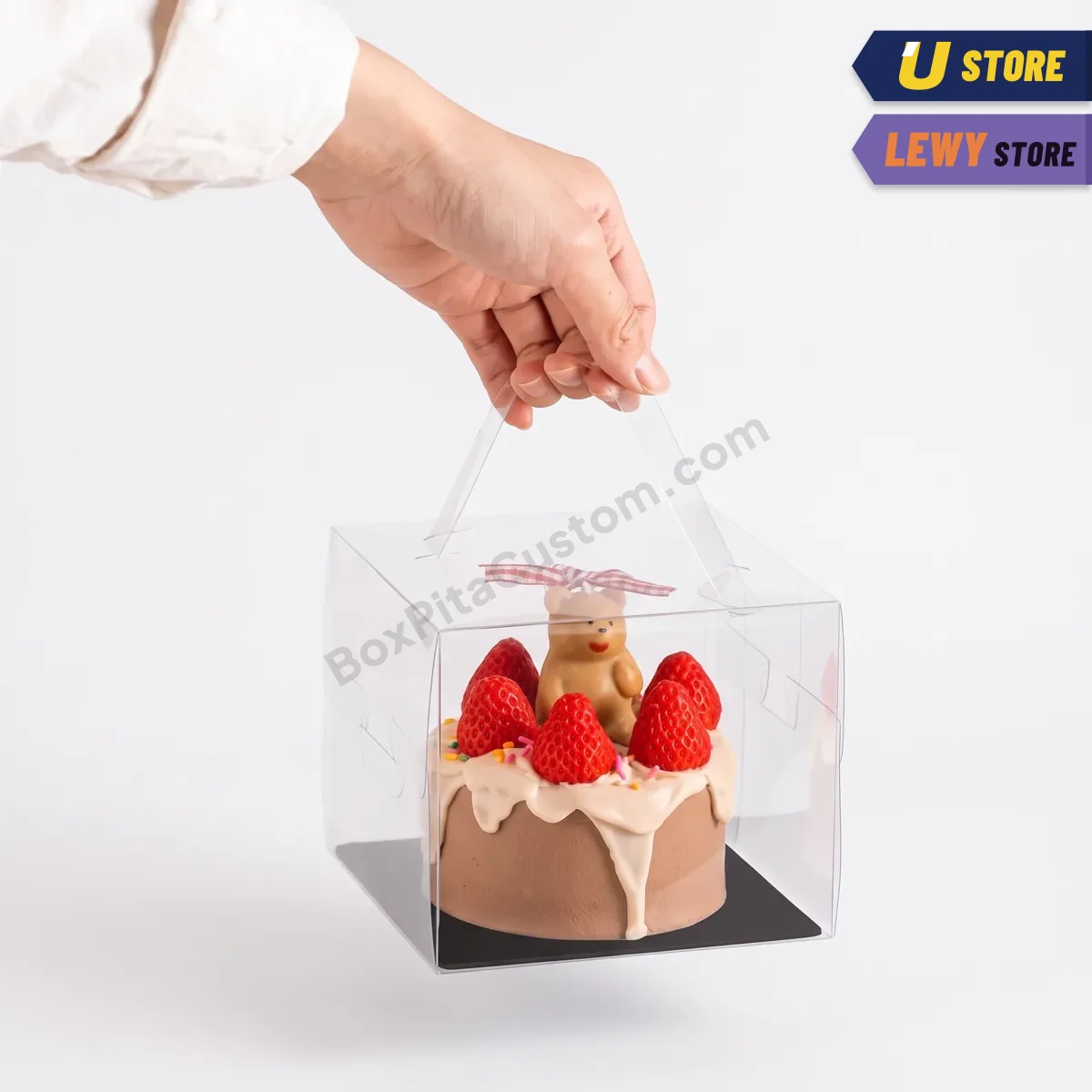 Kotak kue mika bening beralas hitam ditenteng pada pegangannya, berisi kue cokelat berhias stroberi